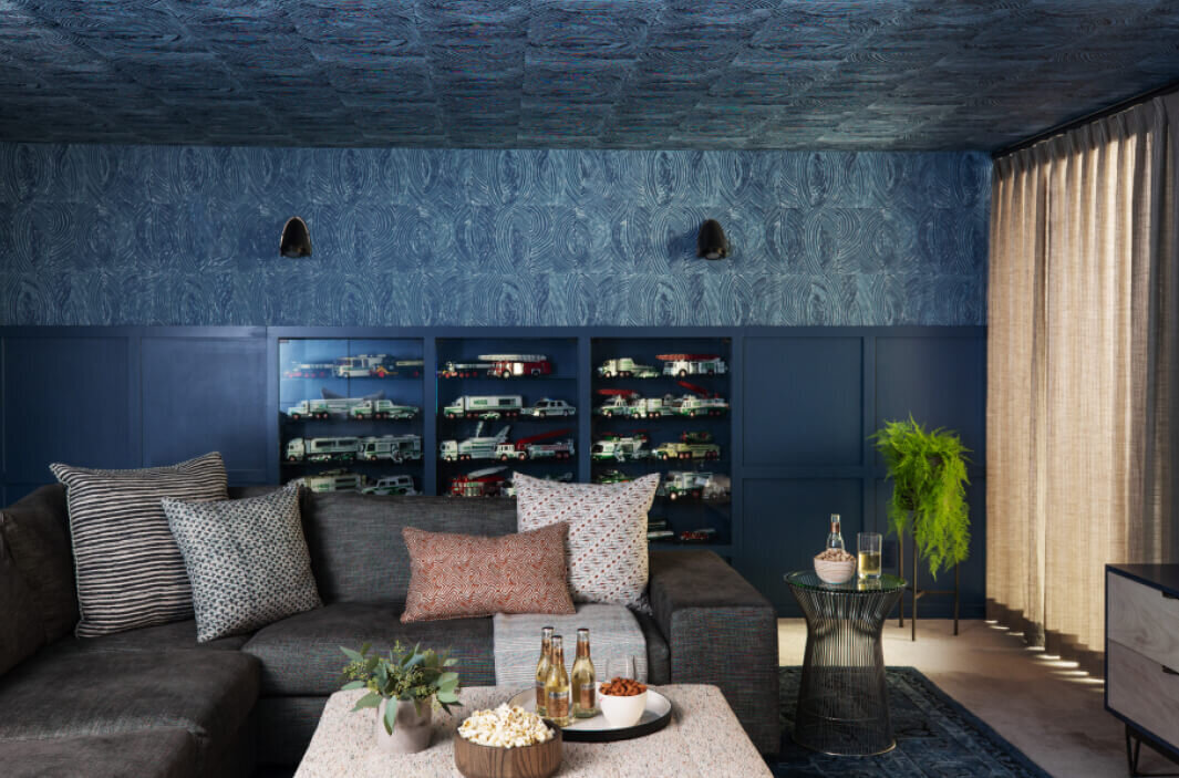 Navy blue media room features large art, blue and green wallpaper on walls and ceiling, blue wainscoting, modern lighting, and custom built-ins for a bold design.
