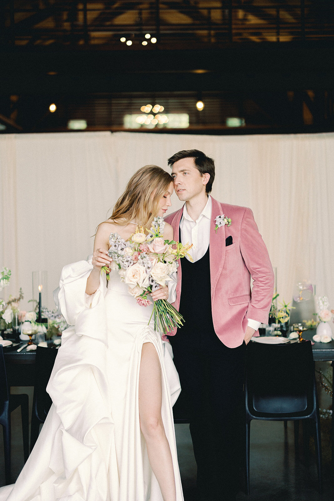 Groom in a pink jacket and bride in an elegant dress holding a pastel colored bouquet
