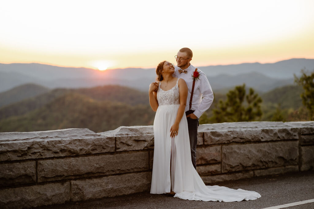 Foothills Parkway wedding photos with bride standing in front of her groom as he holds her shoulders and smiles down at her with the Smoky Mountains in the distance 