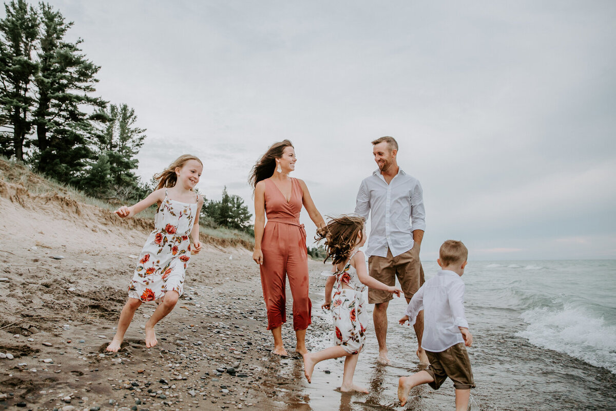 Mom, Dad, and three young children on Grand Bend beach for family photoshoot. Mom and Dad are holding hands walking in the shoreline. Their children, two girls and a boy are running around them barefoot in the water. Mom and dad are smiling at each other.