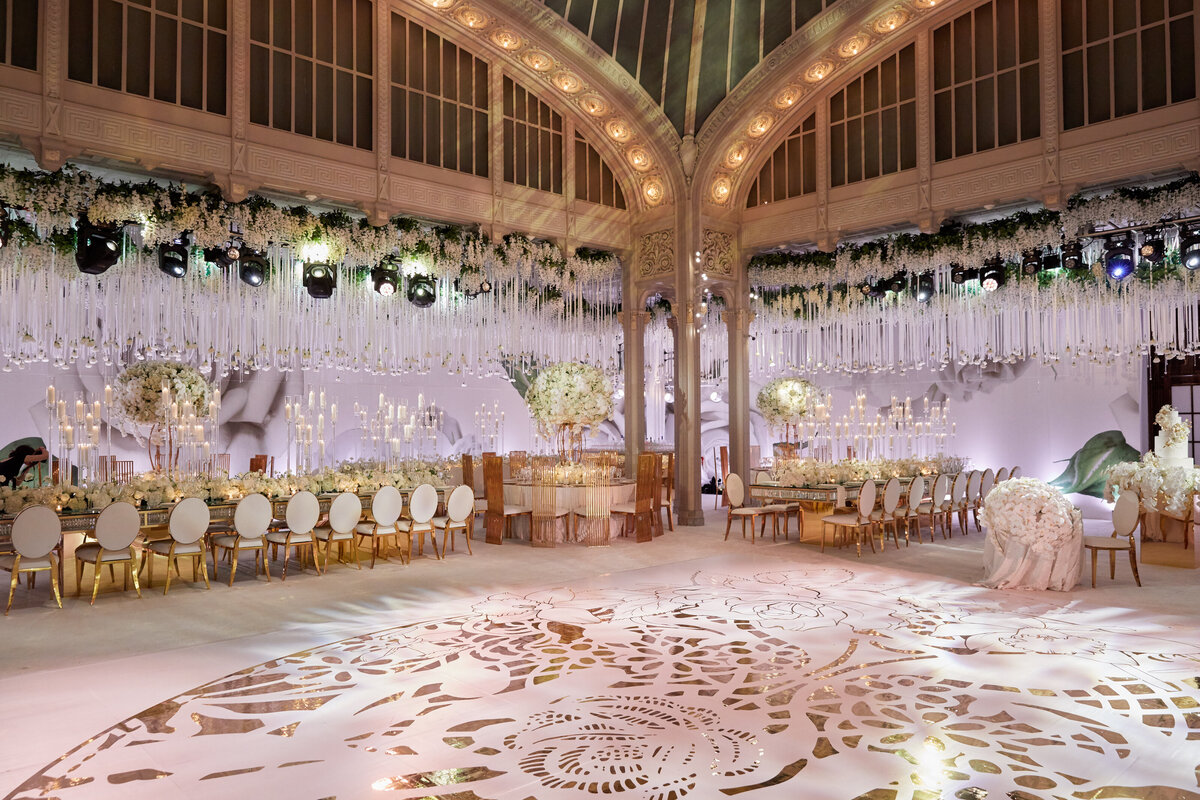The stunning photo was captured at the iconic New York Public Library in New York City by photographer Christian Oth. The lavishly decorated ballroom exudes elegance with its ornate gold and white designs, featuring a mix of round and long tables adorned with exquisite white floral centerpieces and glistening gold tableware. Adding to the grandeur, luxurious hanging white flowers and twinkling lights gracefully cascade from above, casting a romantic ambiance throughout the space beneath the high, arched ceiling. The overall scene captures the essence of opulence and sophistication, making it a truly mesmerizing setting for any grand event.  Furthermore, East West Films provides videography services to capture the beauty and grandeur of events like these. Their expertise in creating stunning visual narratives can complement the photography by Christian Oth, ensuring that every moment is beautifully preserved for years to come.