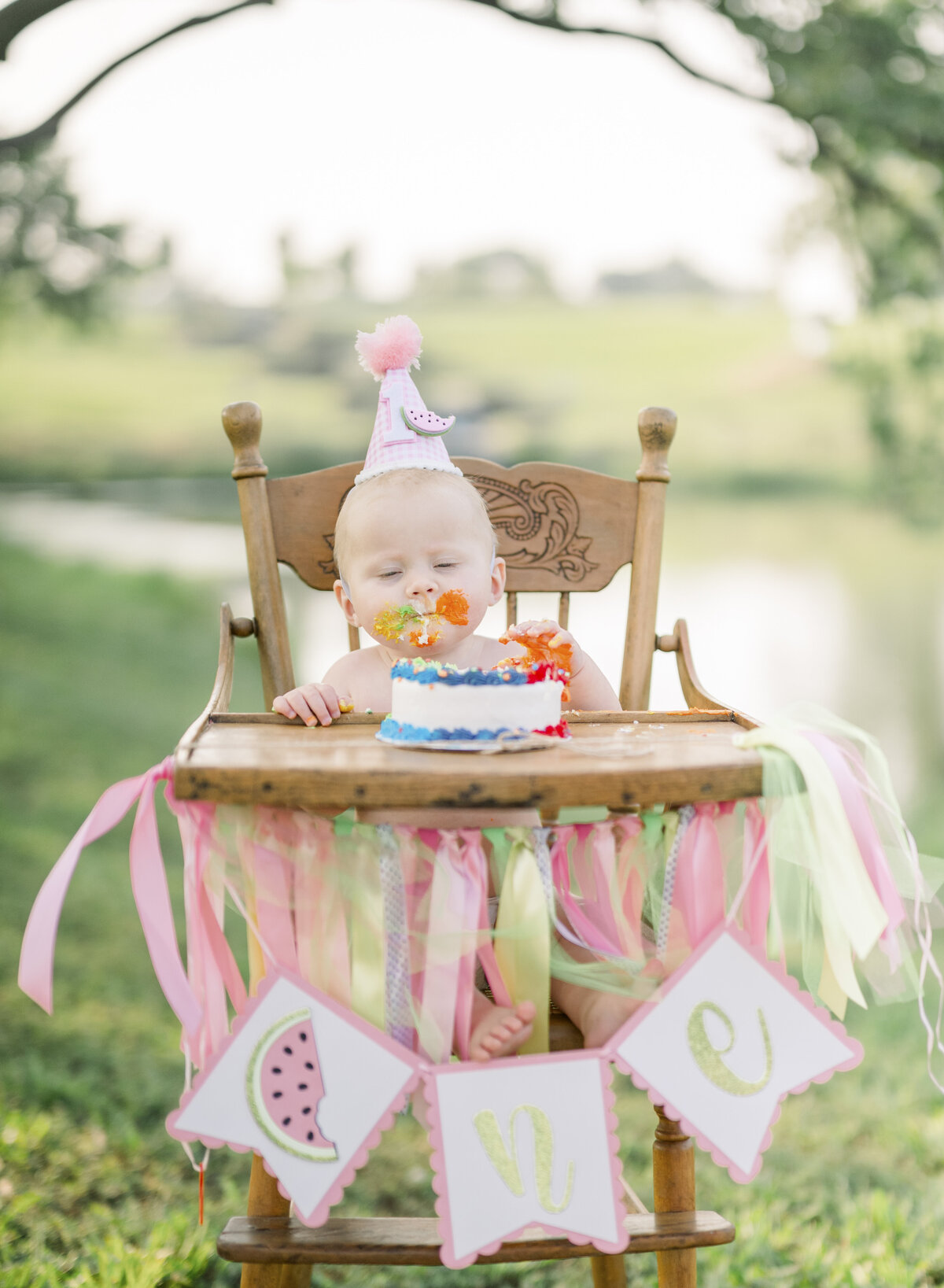 Pink and orange cake smash setup outdoors with balloons and a first birthday banner.