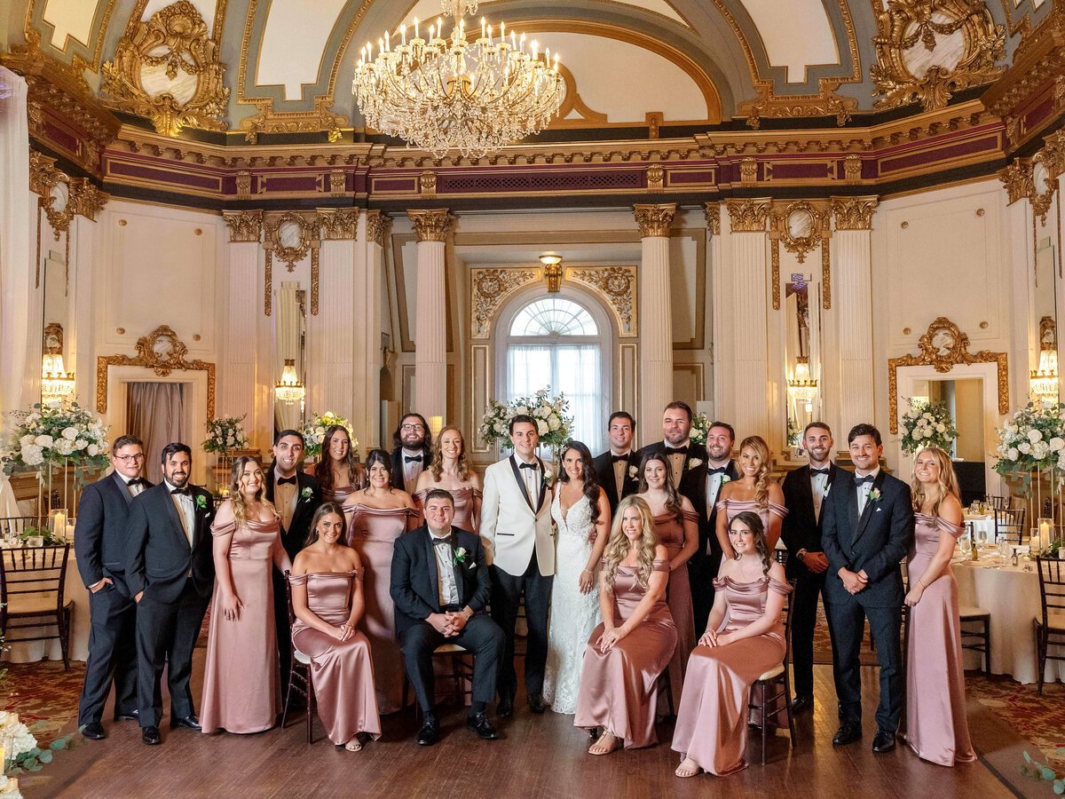 A large wedding party poses in an elegant ballroom adorned with chandeliers and ornate gold details. The group includes a bride in a white gown, groom in a white tuxedo, and attendants in coordinated pink and black formal attire.