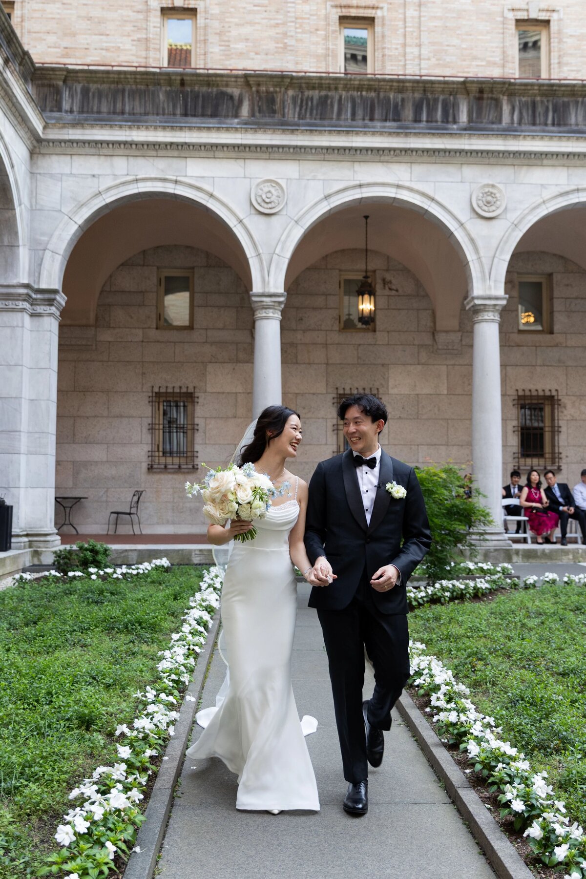 Rhode Island Wedding Photographers | Kelsey Sheehan Photography Timeless Rhode Island Weddings | A joyful couple in wedding attire walk hand in hand through an elegant courtyard with stone arches, surrounded by greenery and flowers.