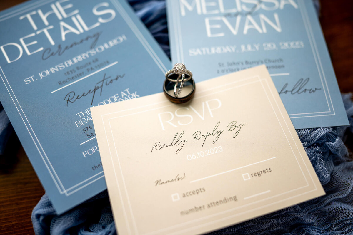 wedding detail photo of the couple's wedding rings set on top of invitations and strips of cloth from bridesmaid's dresses. Captured near window light in Pittsburgh PA.