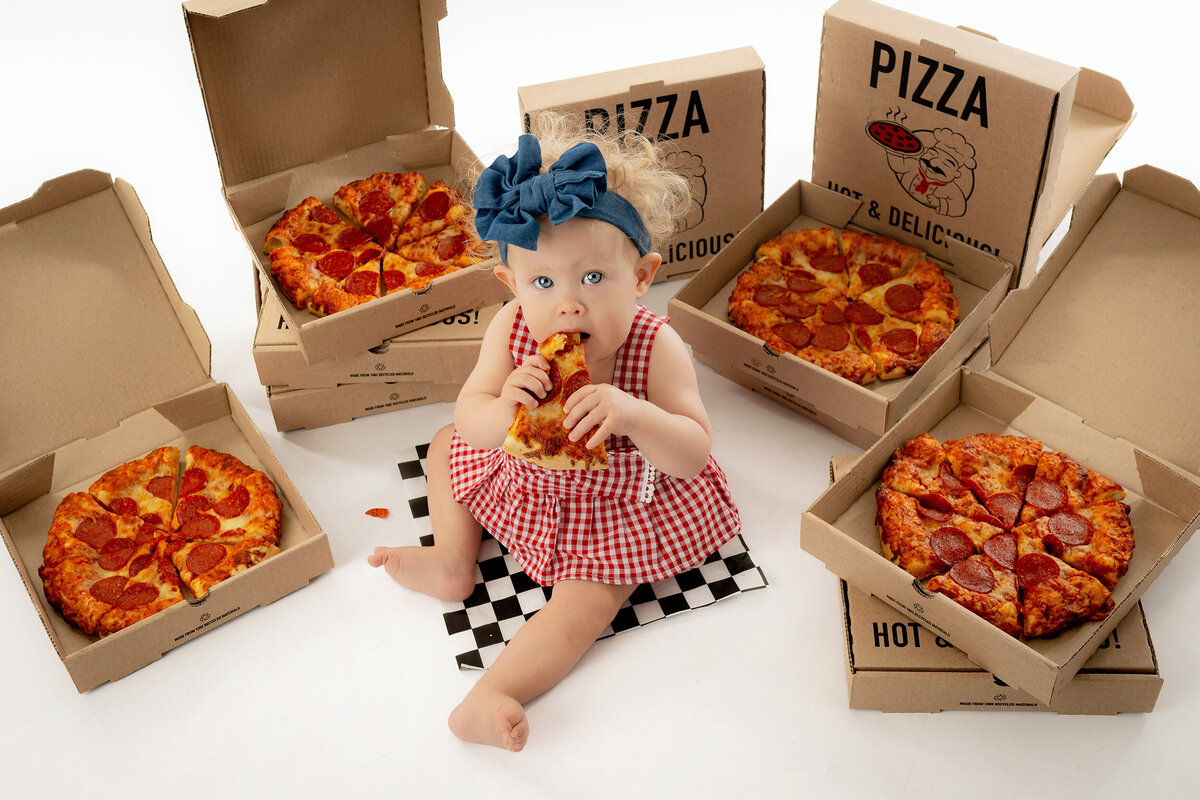 Little girl surrounded by pizza boxes sits and eats a slice.