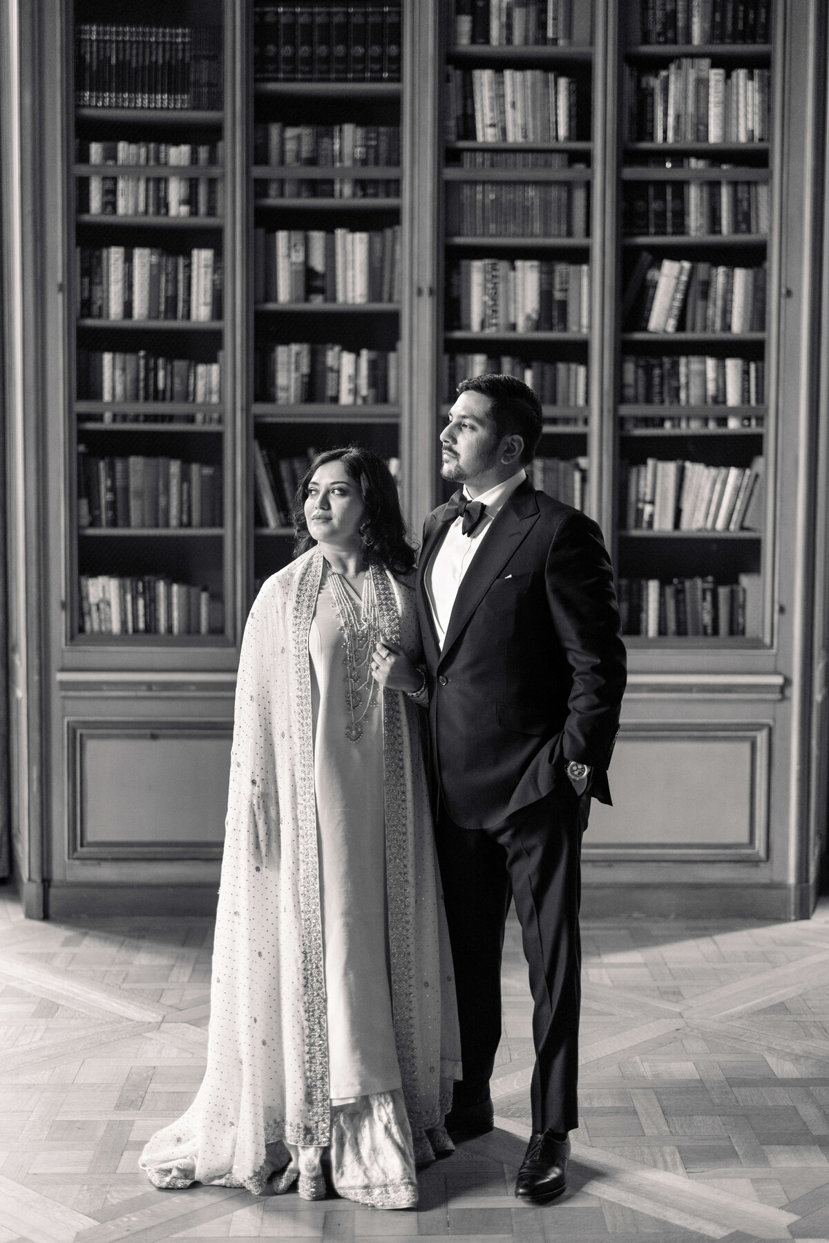 A couple stands elegantly in front of a tall bookshelf, their attention directed upward. The woman wears a traditional outfit with a cape, while the man is in a suit and bow tie. The scene is in black and white, with a wooden floor adding warmth.