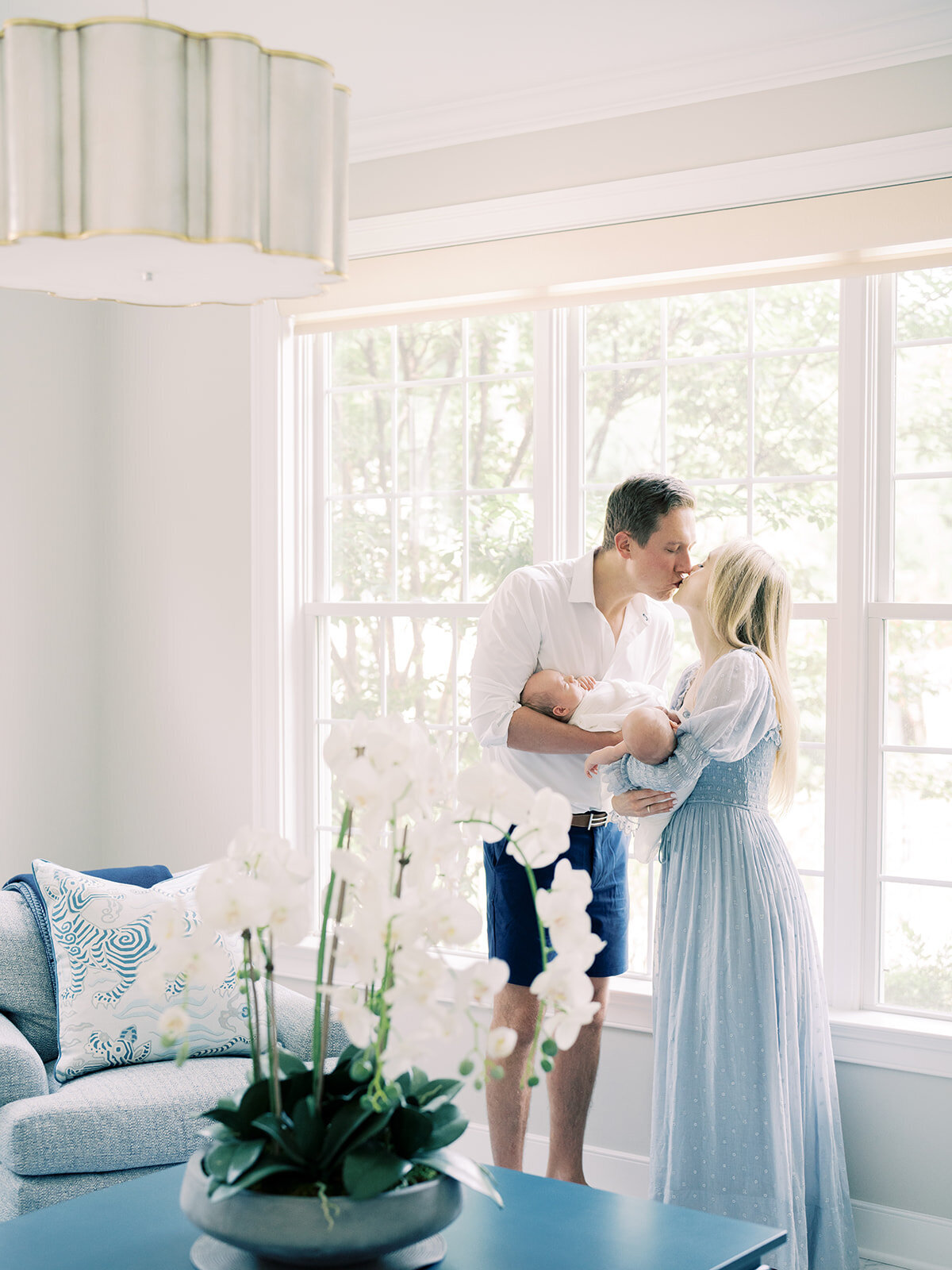 Mother and father lean over to kiss while holding their twin baby boys in their living room by the window.