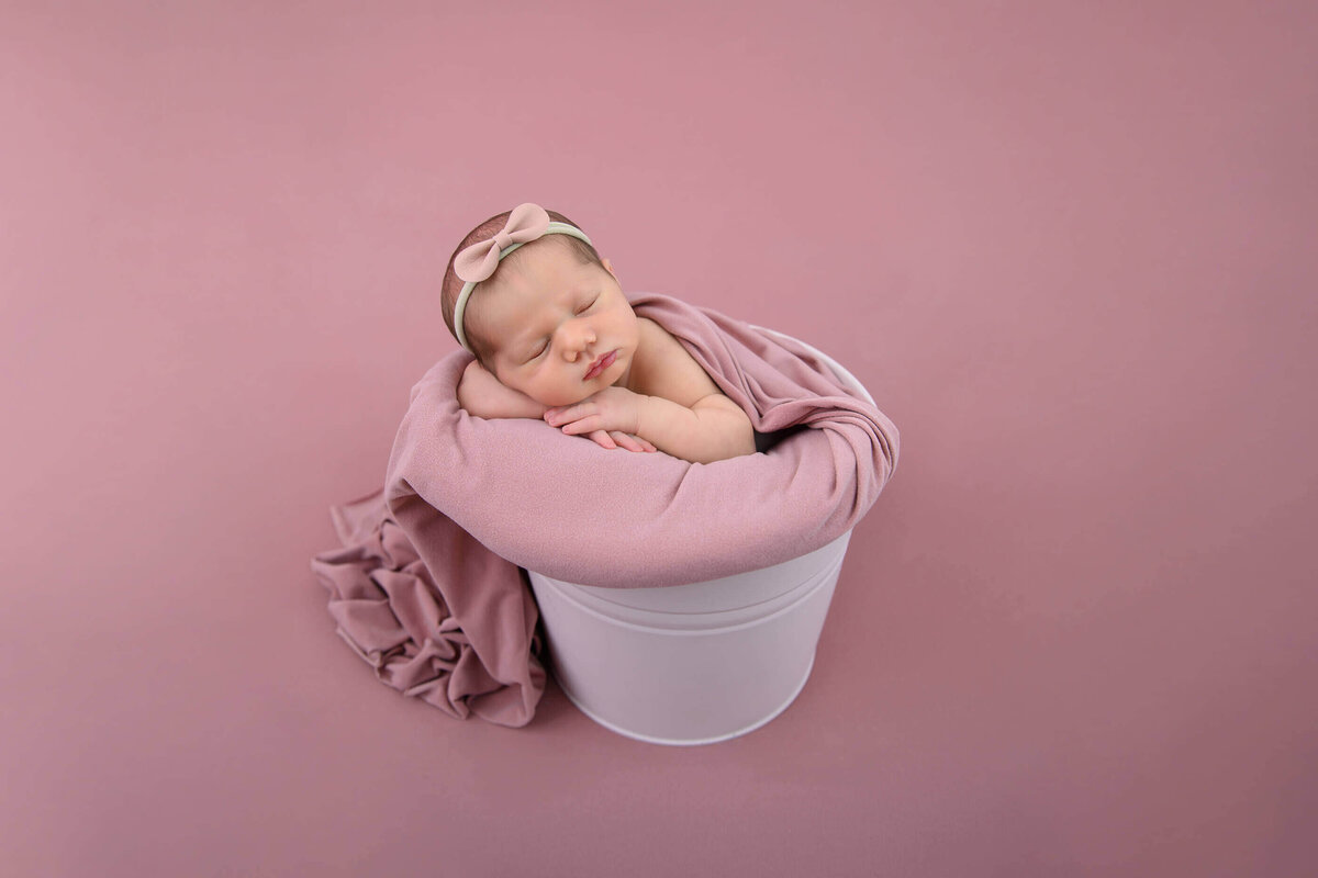 newborn baby girl in a pink bucket on a pink background for her Burlington newborn photography session.