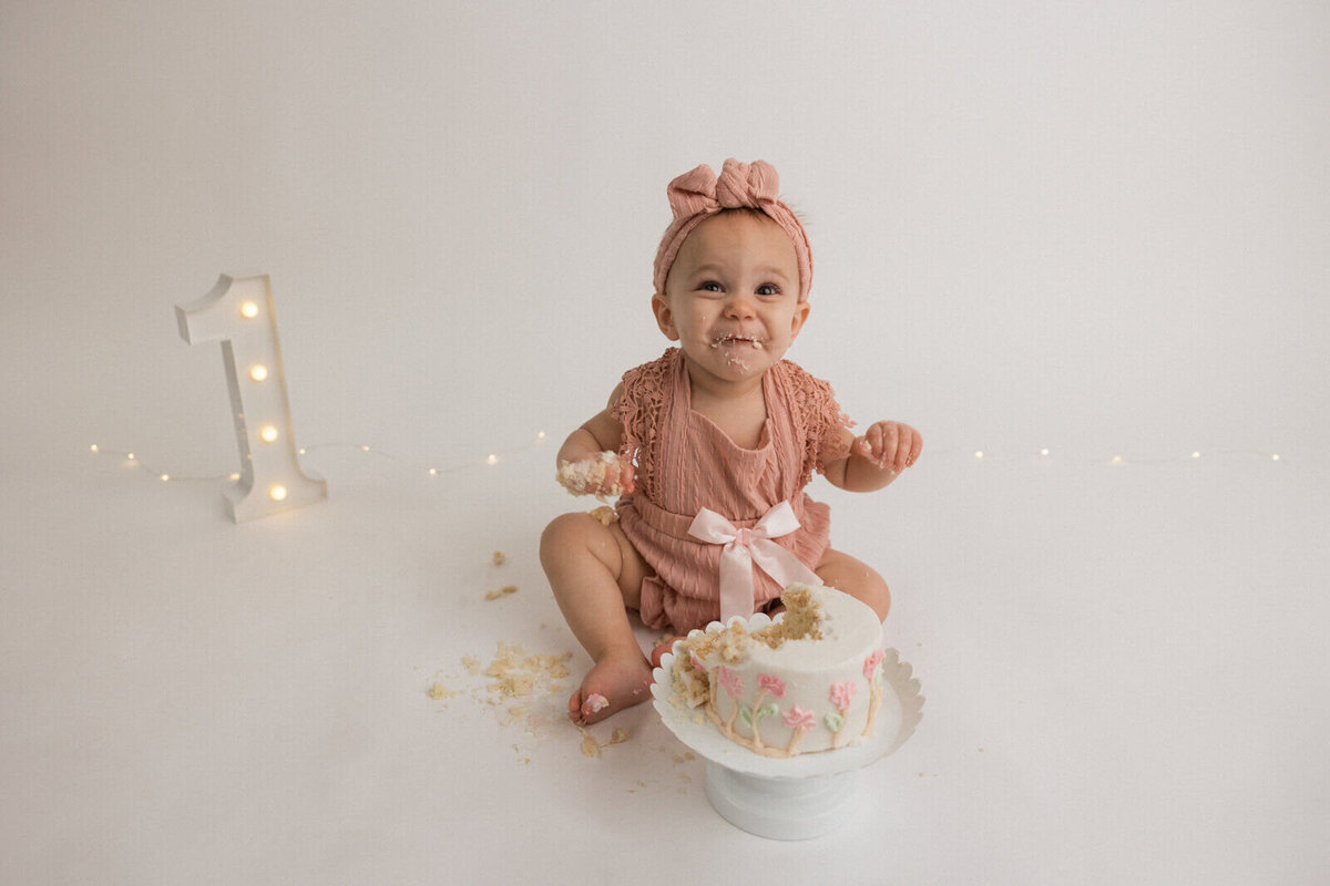 A baby girl in a pink romper and headband sits next to a half-eaten cake decorated with pink flowers. She has cake on her hands and face and a lit number one sign is in the background.