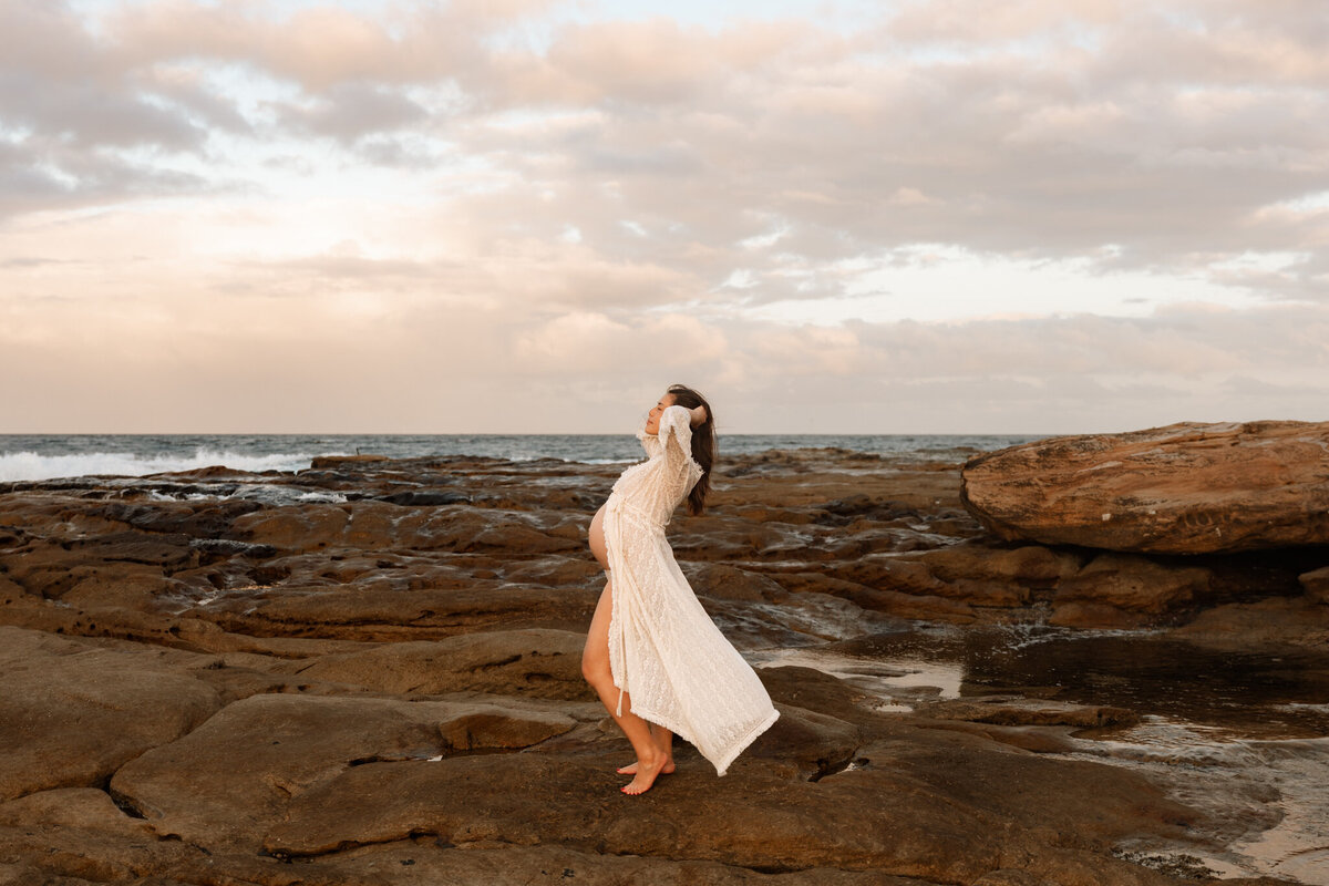 Pregnant woman in a sheer lace robe posing on coastal rocks at sunset during a Sydney beach maternity photography session.