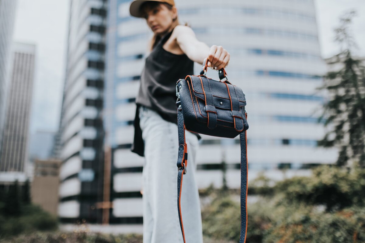 Woman in jeans and a cap stands in front of skyscraper holding a black purse in front of the camera.
