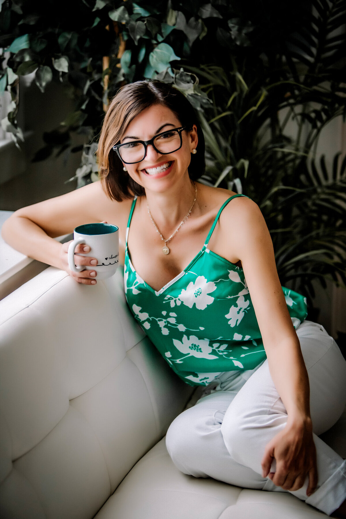 A female dating portrait client wearing a green shirt with white flowers, sitting on a white couch, holding a cup of coffee & smiling at the camera
