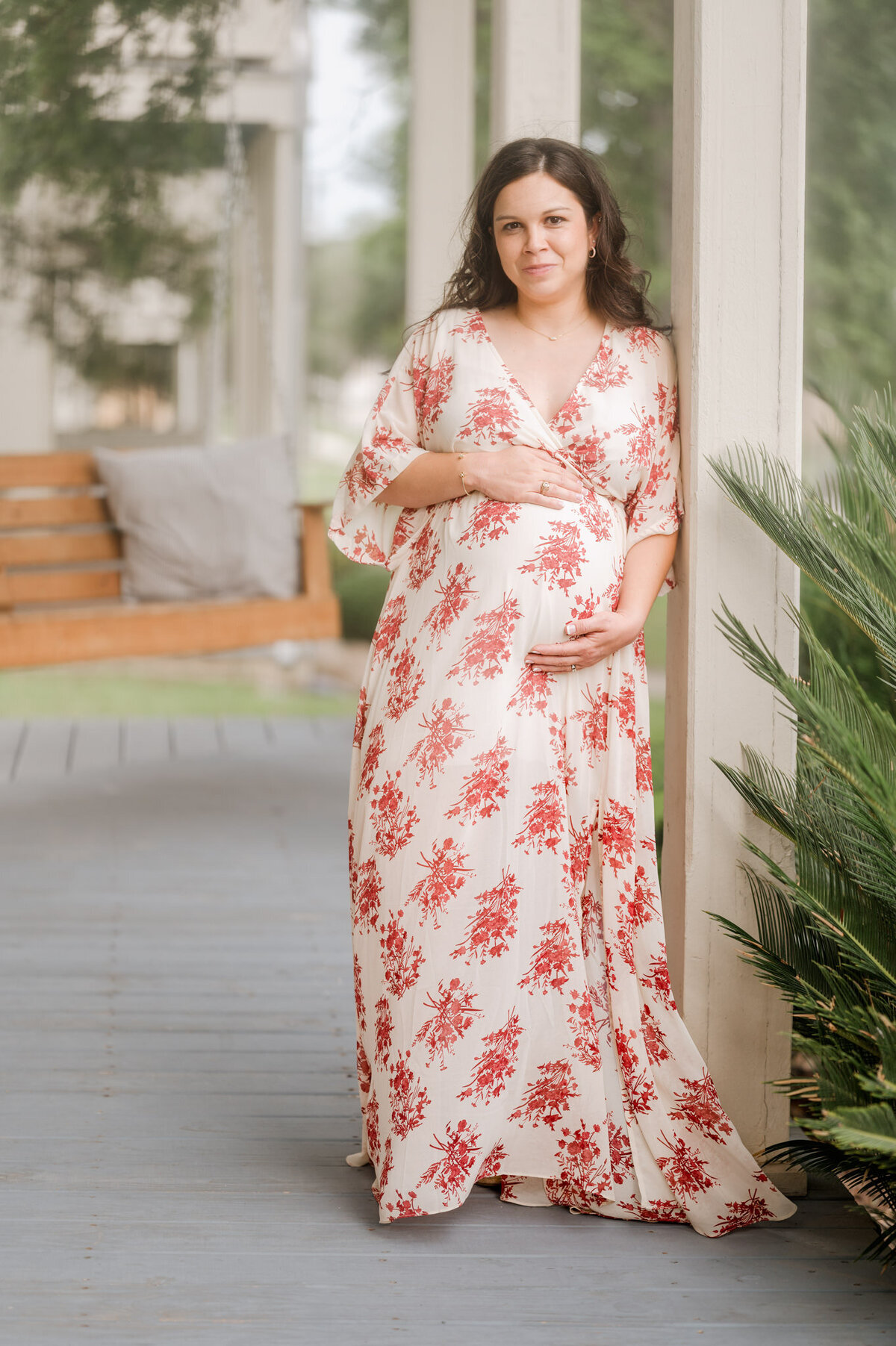 Portrait of an expecting woman in a floral gown leaning against the column of a porch.