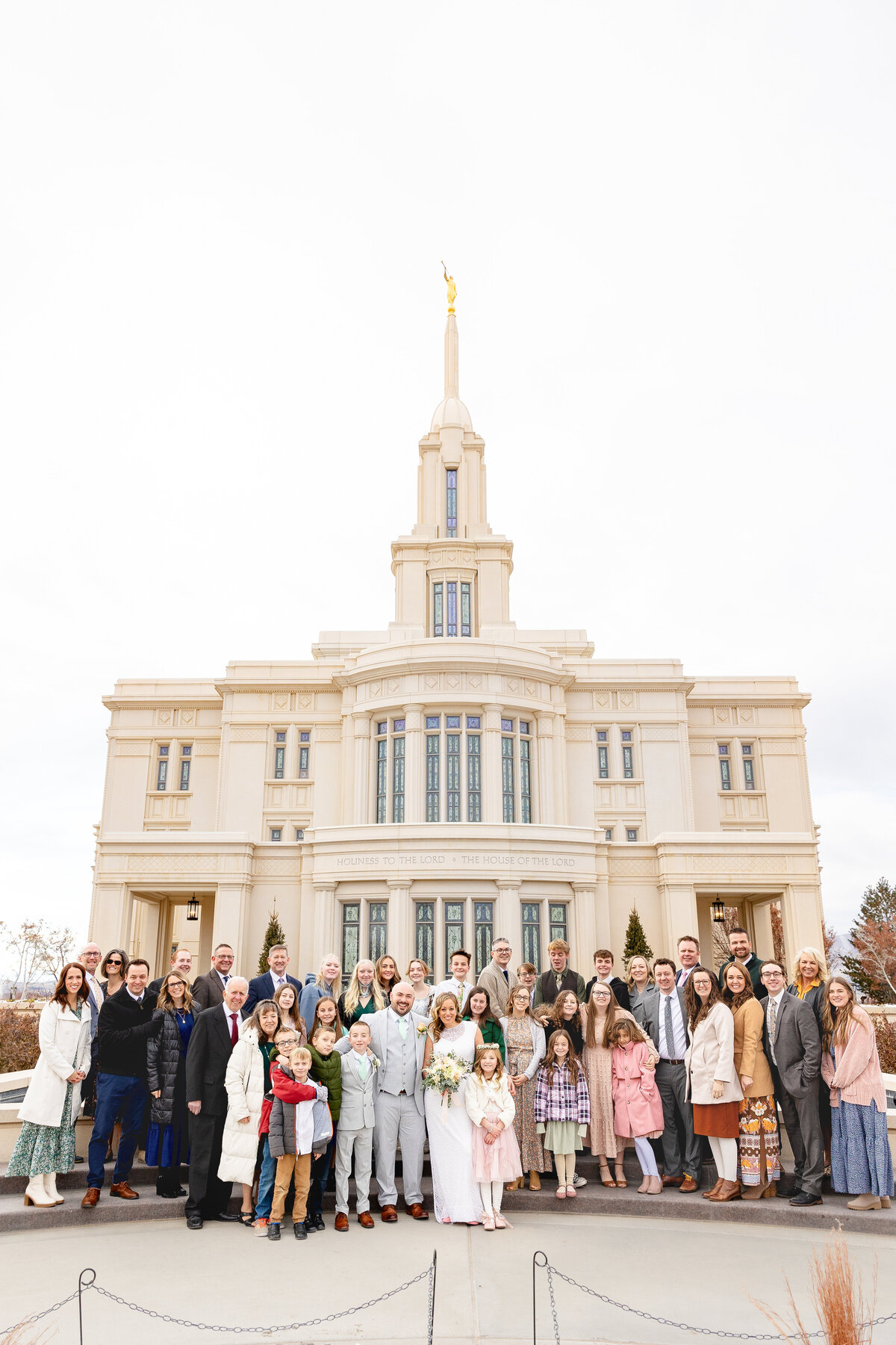 A large wedding group gathers on the steps of the Payson Utah LDS Temple for a formal photo, all dressed warmly for the season with smiles and excitement.
