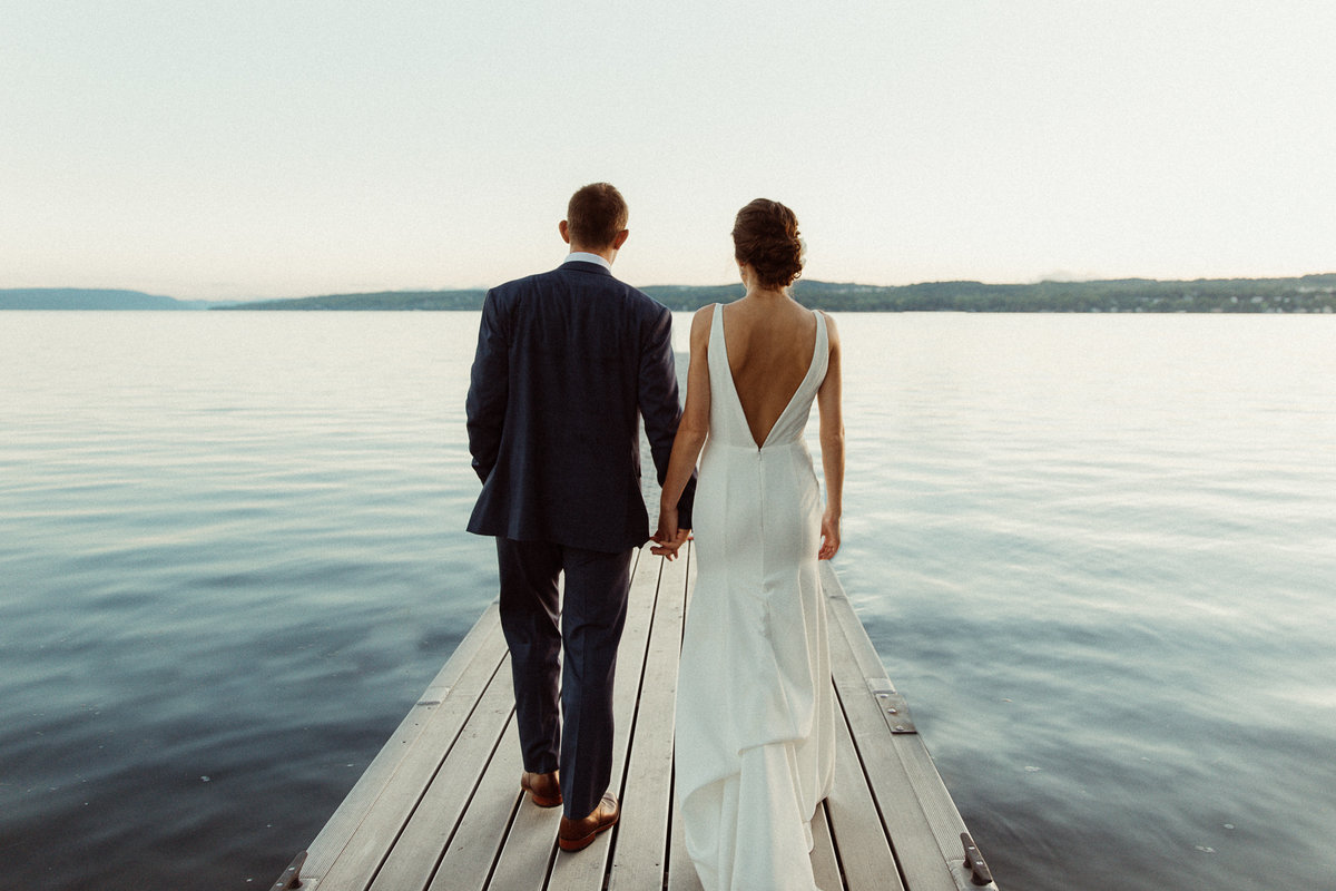 bride and groom holding hands, walking on dock towards Canandaigua Lake