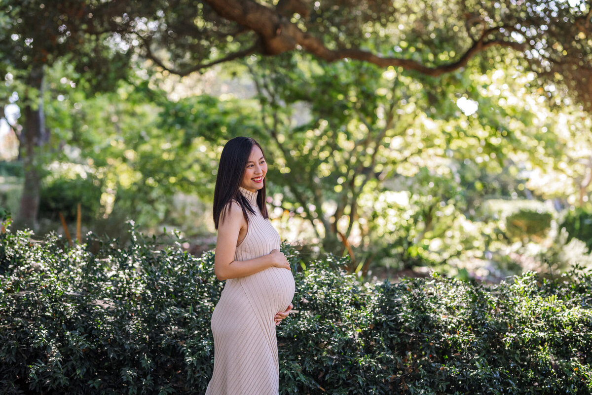 pregnant woman in off-white dress smiling at someone off camera and holding her belly in Berkeley gardens