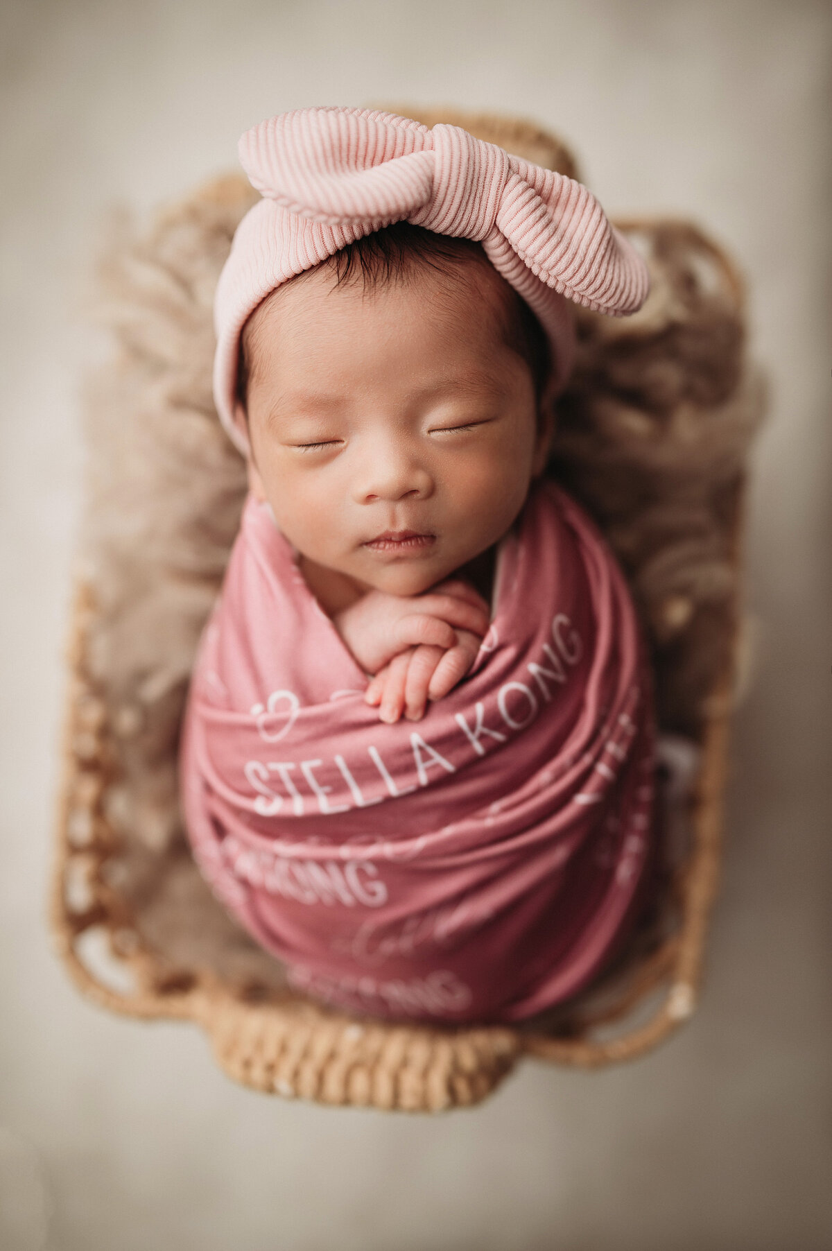 sleeping newborn baby girl  swaddled in personalized name blanket and wearing a big pink bow laying in a layered  tan basket  with a cream background