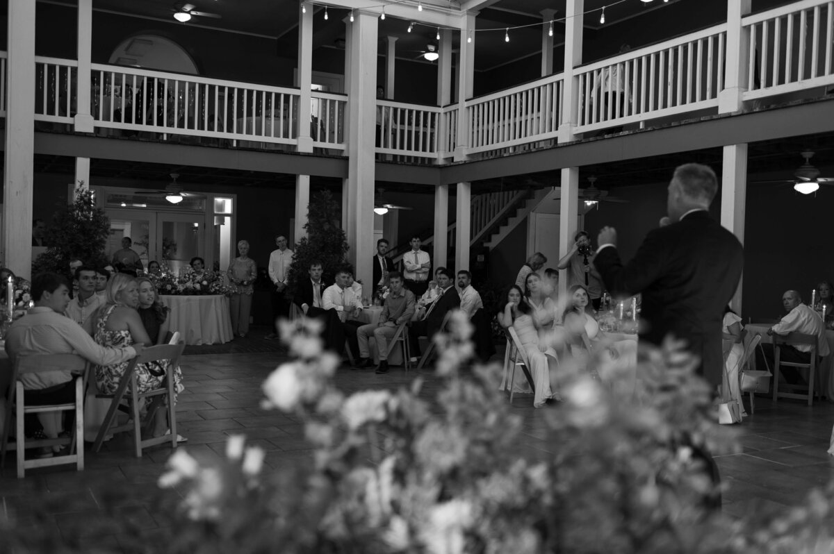 A man in a suit speaks to a seated audience at an outdoor wedding reception, held in a two-story venue with string lights and floral decorations.