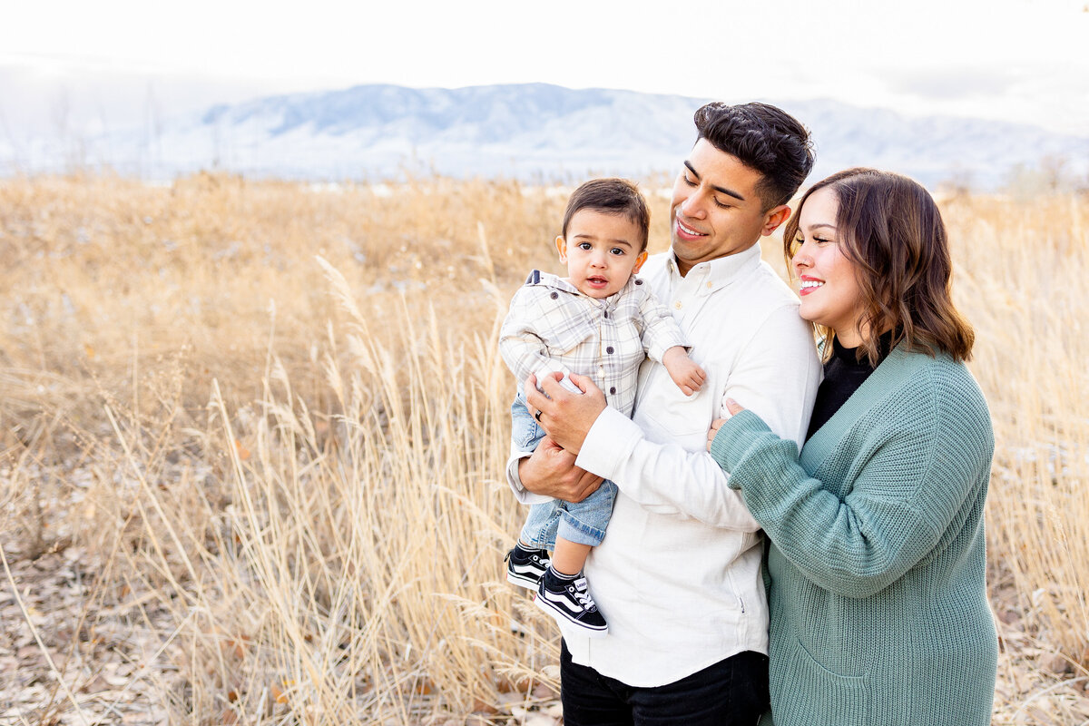 A father in a cream-colored shirt cradles his young child in his arms while the mother, dressed in a green sweater, leans in with a joyful smile, set against a backdrop of golden fields and distant mountains.
