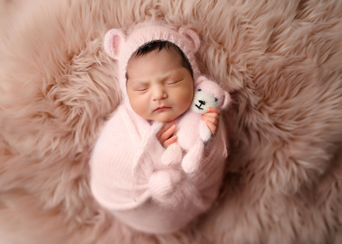 newborn girl swaddled in angora wrap wearing a pink bear bonnet and holding a tiny bear on a shaggy pink rug