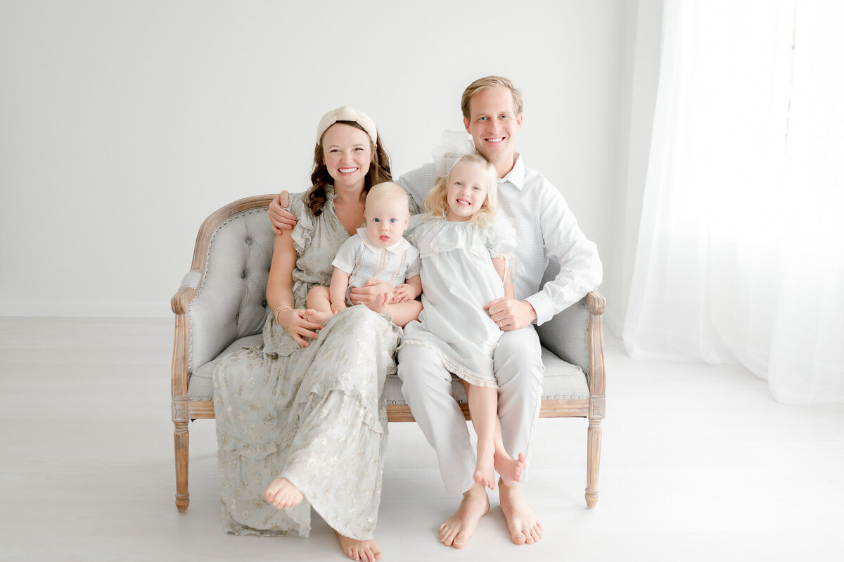 Family of 4 sit on a gray bench in a white studio