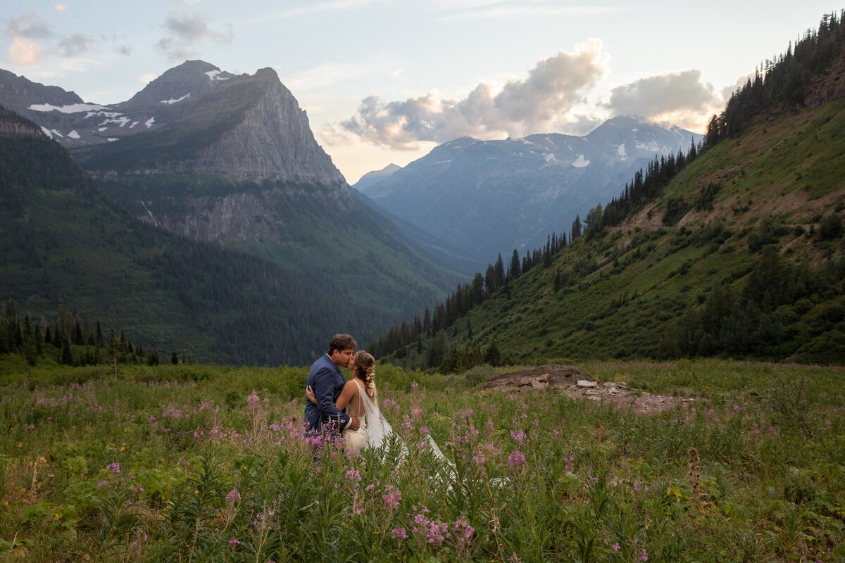 A bride and groom stand kissing in a meadow full of wildflowers in Glacier National Park on their elopement day.