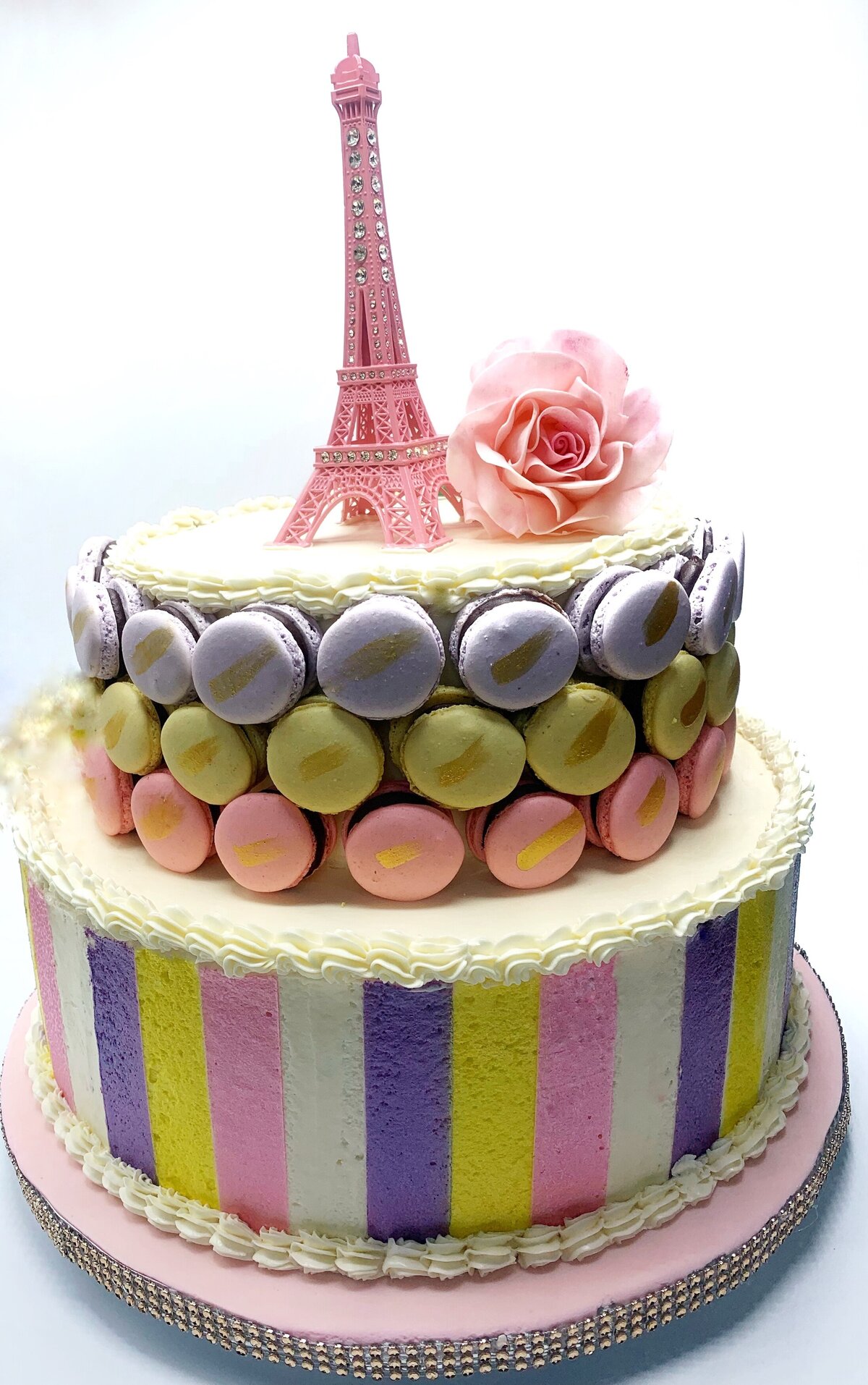 Paris inspired 2 tier buttercream cake  decorated with pink, purple, yellow stripes , colored macarons. Eiffel tower and handcrafted sugar flower rose on top