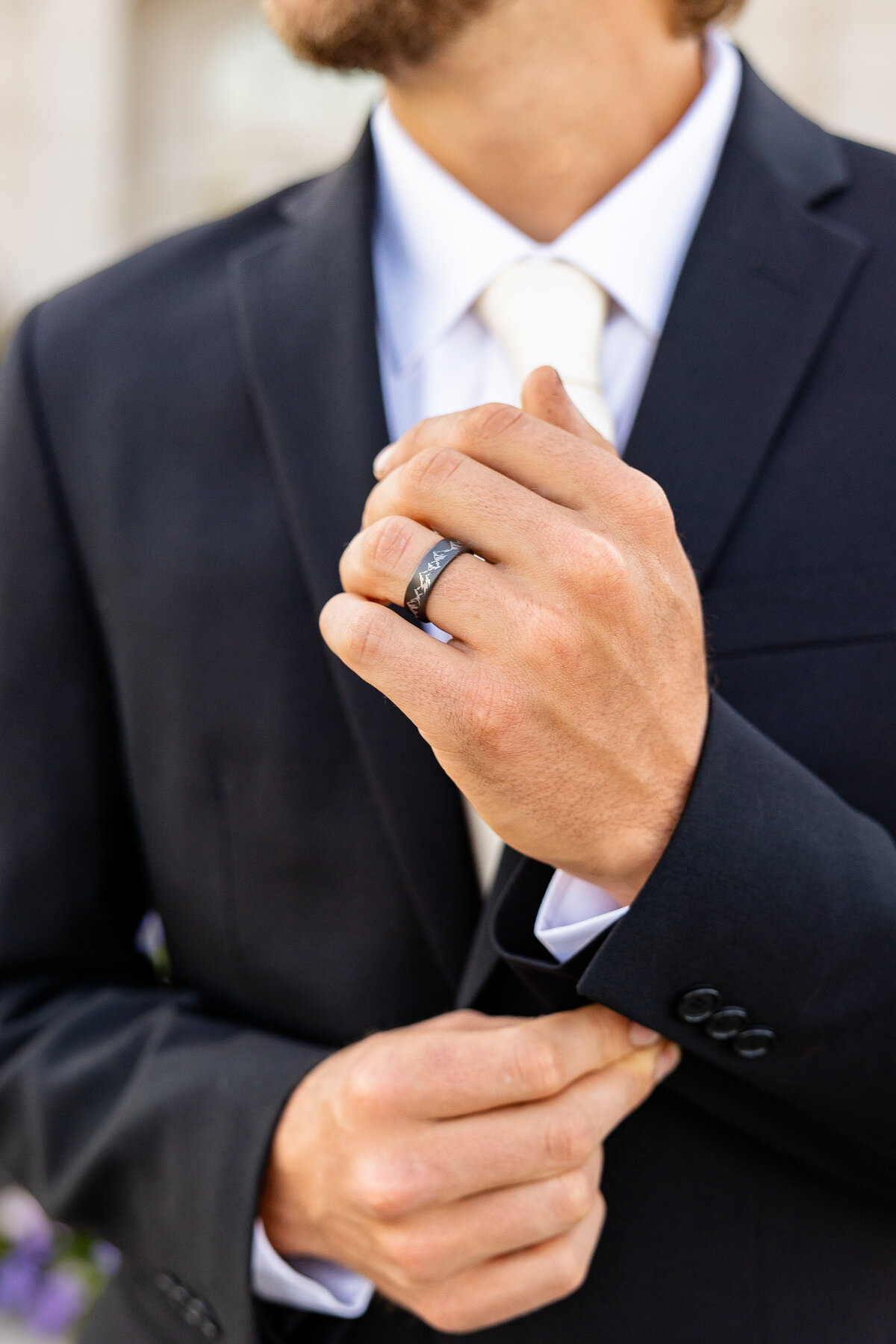 A groom in a black suit adjusts his jacket sleeve, showing off a matte black wedding ring with mountain engraving