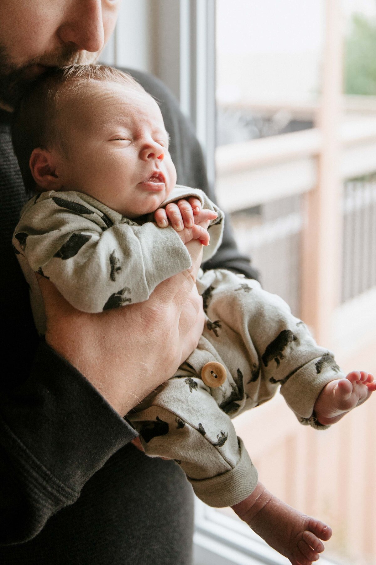 In-home newborn session - Dad holding baby against his chest baby facing outward toward the window. Baby curled up in a onesie