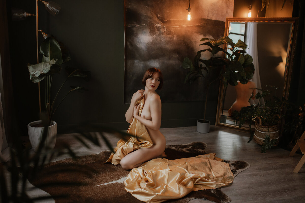A nude woman holds a gold sheet as she poses on the floor of the Victoria, BC boudoir studio.