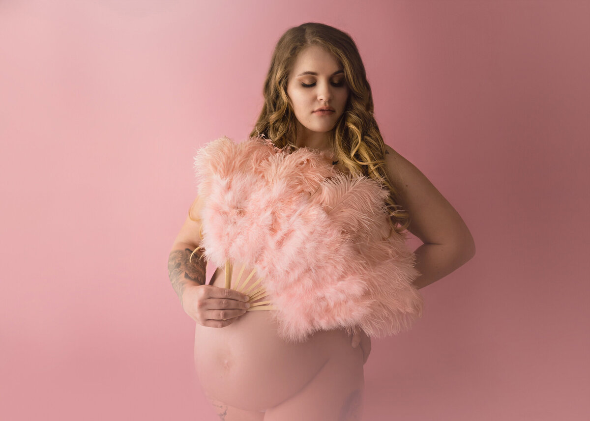 A pregnant woman stands in a studio covering with a pink feather fan while looking down her shoulder in a pink studio of an Iowa maternity photographer