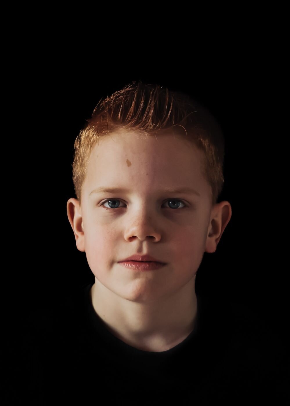 Een portretfoto van een jonge jongen met een open en vriendelijke uitstraling, gefotografeerd tegen een donkere achtergrond