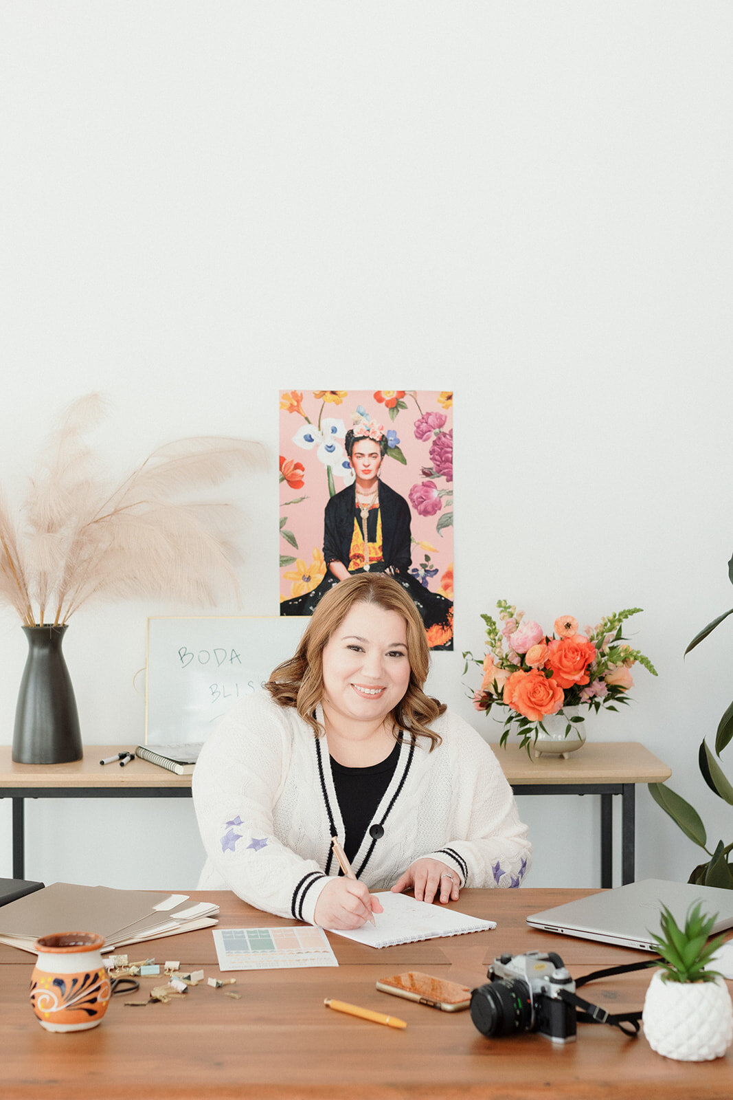 casual business owner sitting at her desk and filling out papers while smiling with Frida Kahlo poster behind her