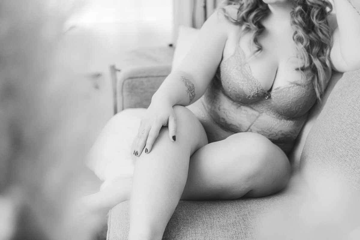A close-up image of a woman wearing gray lace lingerie, with her tattooed arm gently resting on her leg as she lounges on a couch.