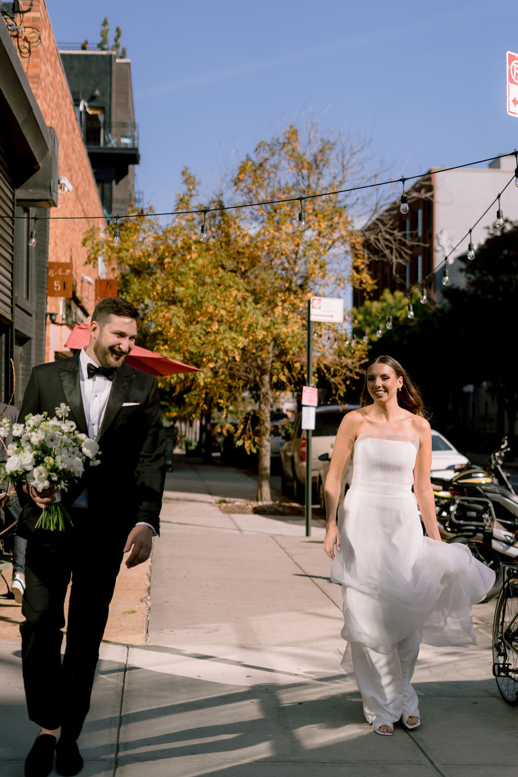 married couple walking down a sidewalk while the groom holds a bouquet
