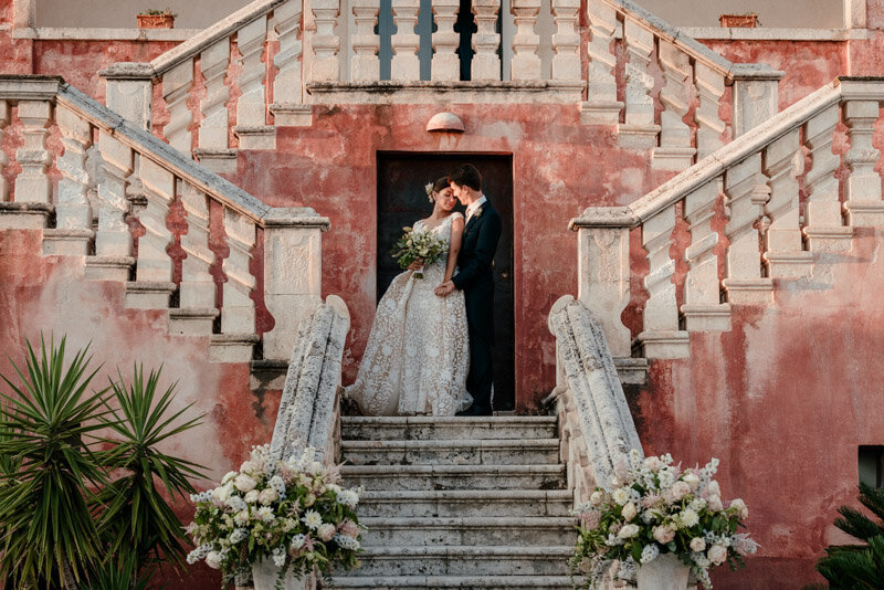 Mariés posant avec élégance sur un escalier en pierre devant une façade rose, lors d’un mariage romantique dans les Pouilles près de Monopoli, en Italie.