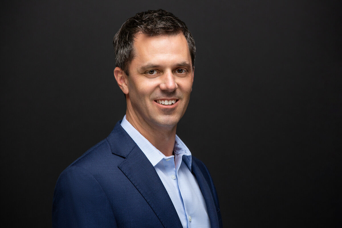 A businessman in a blue suit poses for a headshot on a dark grey background for Janel Lee Photography studios in Cincinnati Ohio