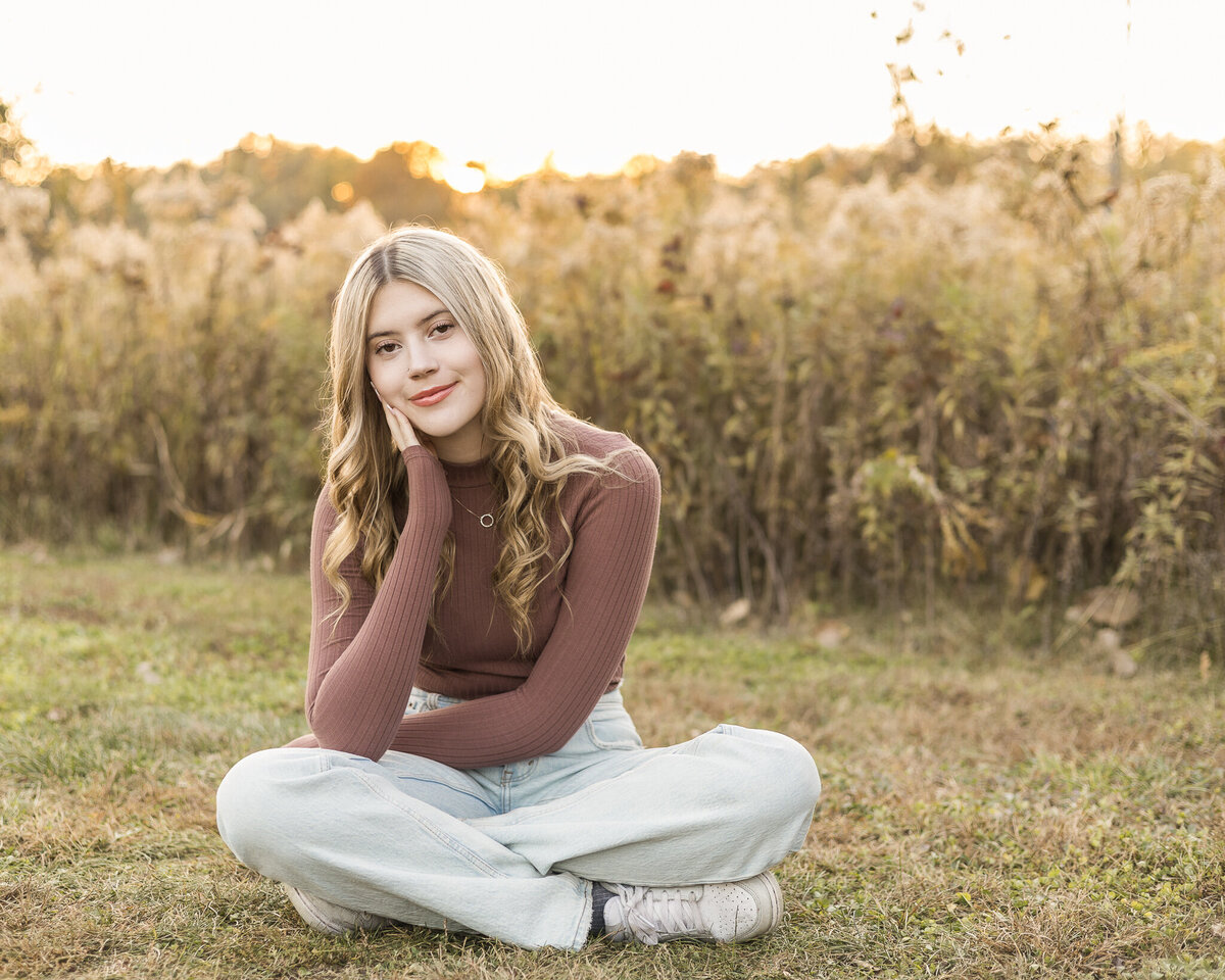 High school senior female with long blonde hair, sitting in the grass, with trees in the background, wearing a long sleeve purple shirt, blue jeans, looking at the camera with a soft smile