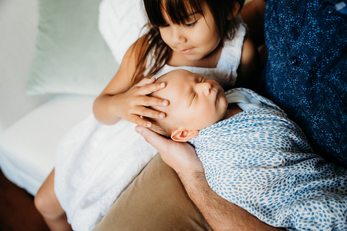 Newborn Photographer, a dad holds his baby as older sister looks on with hand tenderly touching baby's head