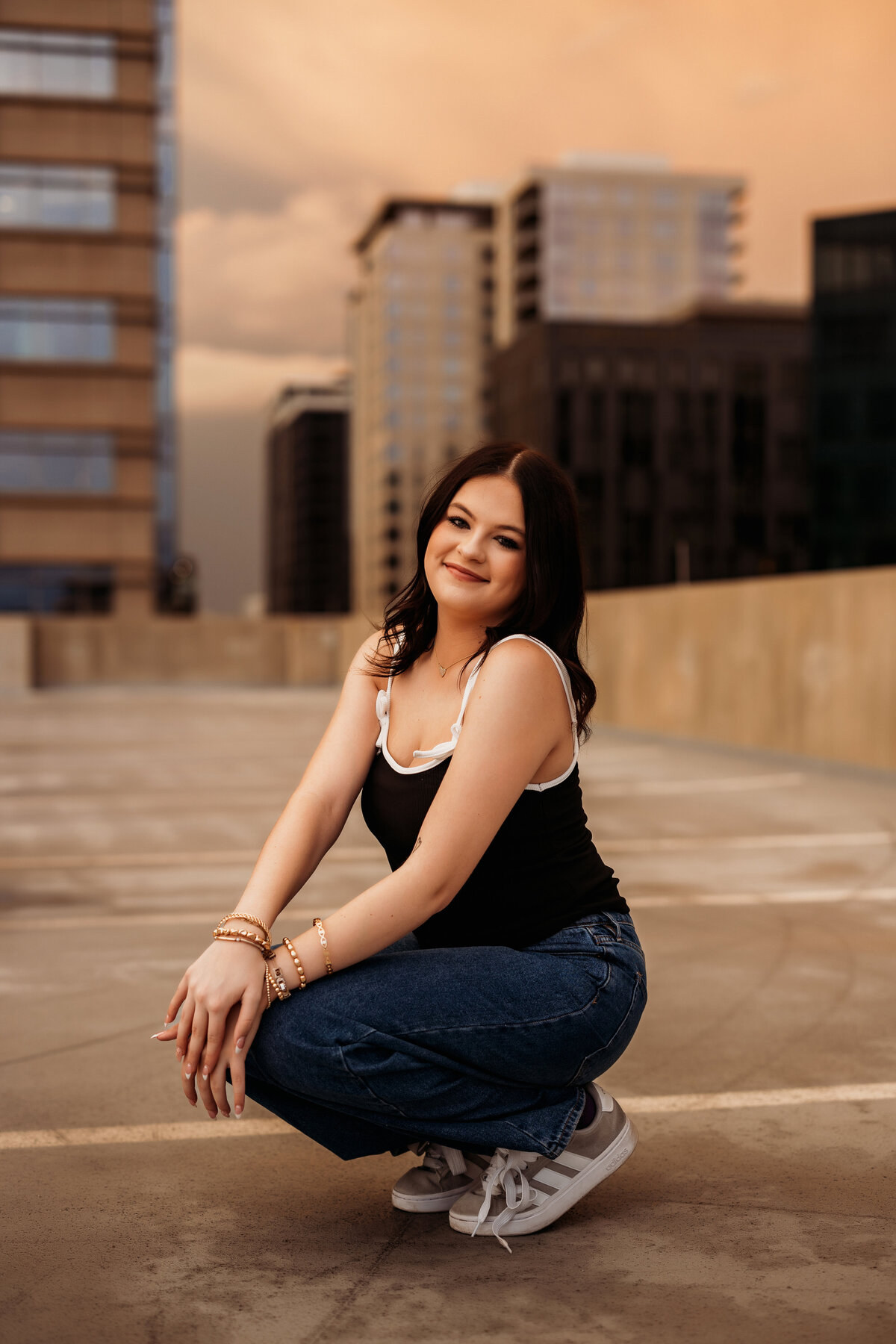 high school senior girl squats on a rooftop for an urban senior photo session with large buildings in the background