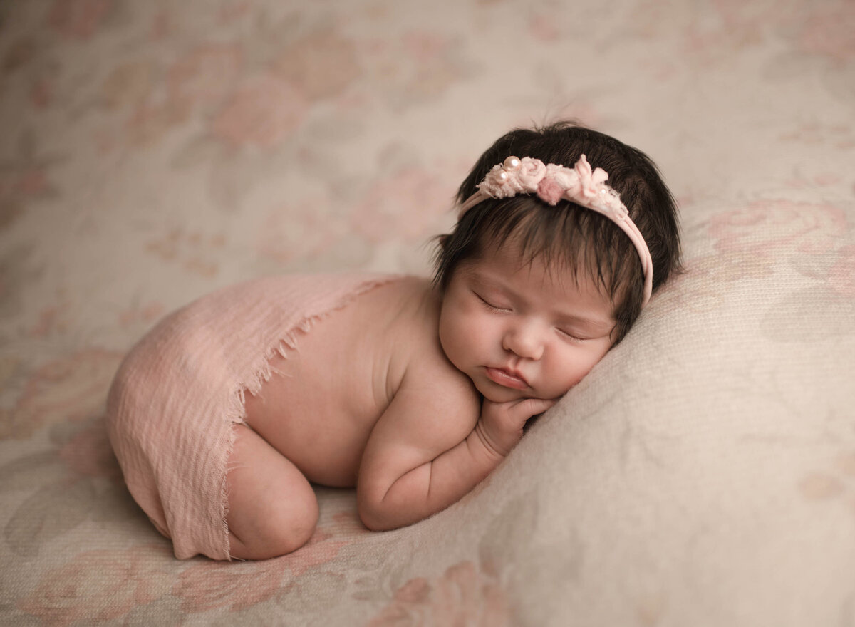 Side profile image. Menifee newborn photoshoot. Baby girl is sleeping on her belly with her hands under her chin. Her face is positioned so it is facing the camera. Baby has blush fabric draped atop of her.  Captured by best Menifee newborn photographer Bonny Lynn Photography