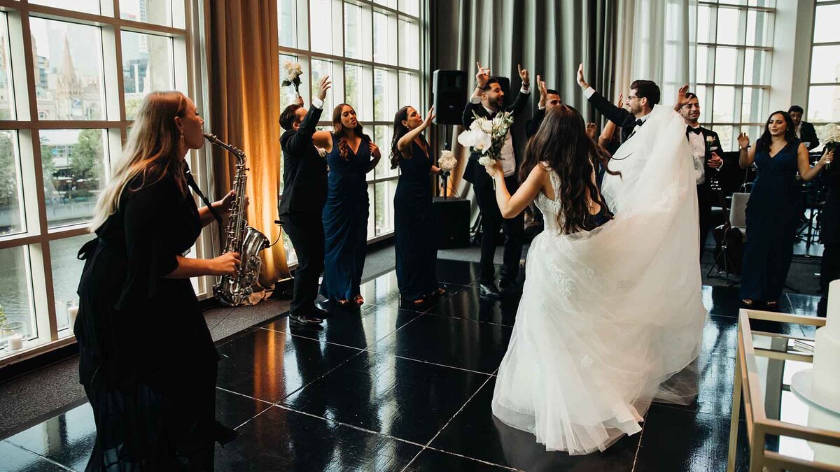 Bride, bridal party, and guests dancing to a live saxophonist at an elegant indoor wedding celebration