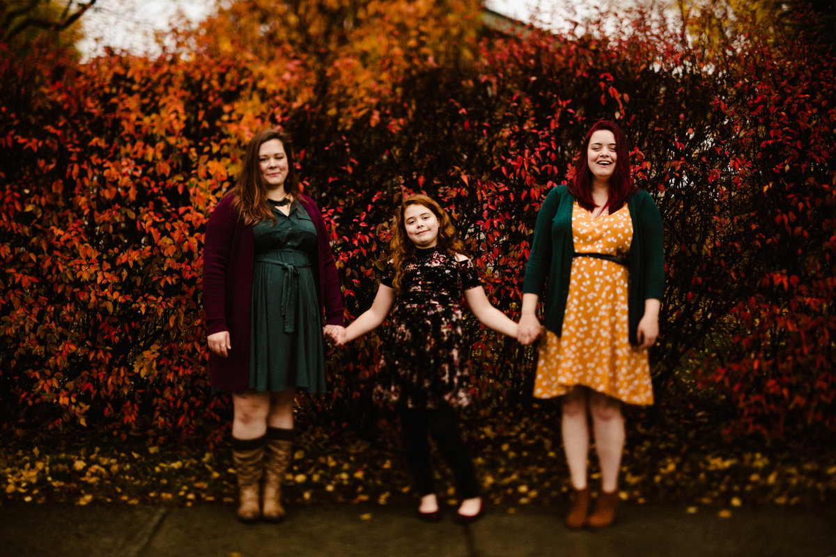 mom and two daughters wearing colorful dresses stand in front of vibrant red bushes