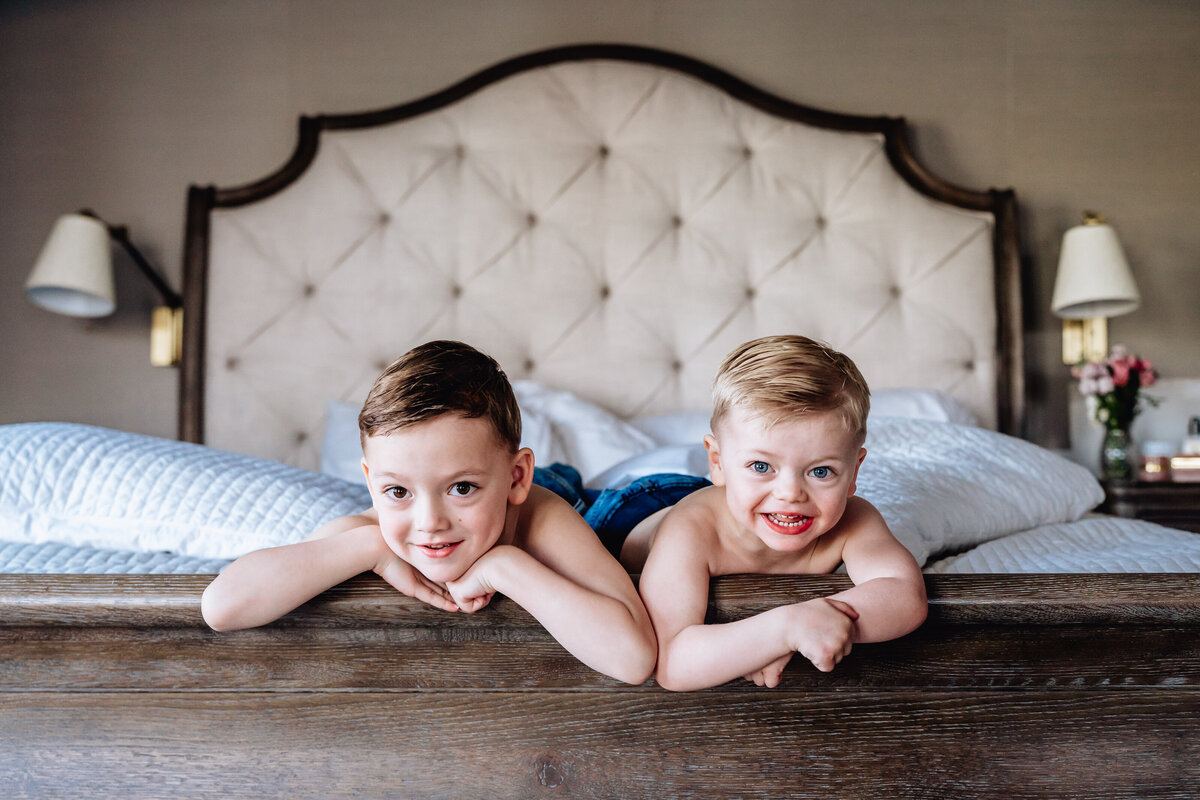 Two young boys lie on their stomachs on a bed, resting their chins in their hands and smiling mischievously at the camera.