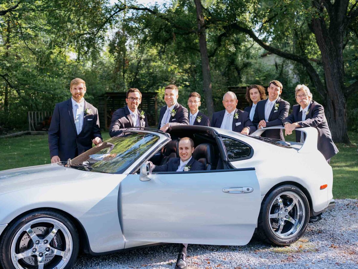 Groom in sports car surrounded by groomsmen.
