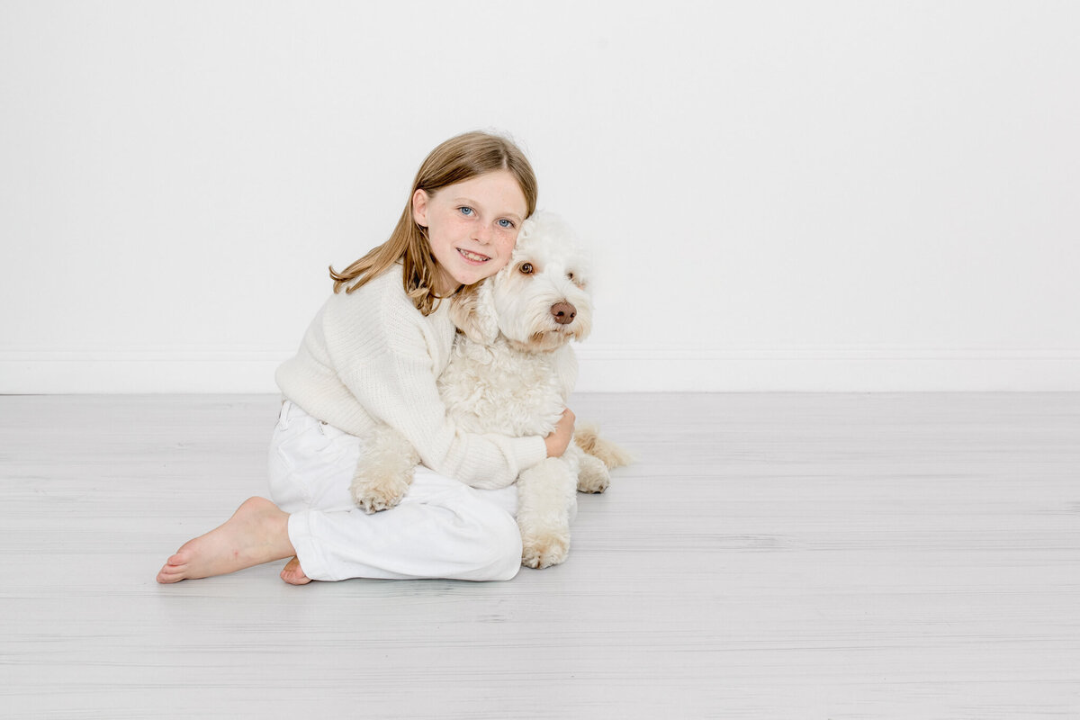 A young girl with shoulder length blonde hair wearing all white, hugging her big, white dog while sitting on the ground and smiling at the camera in a studio.