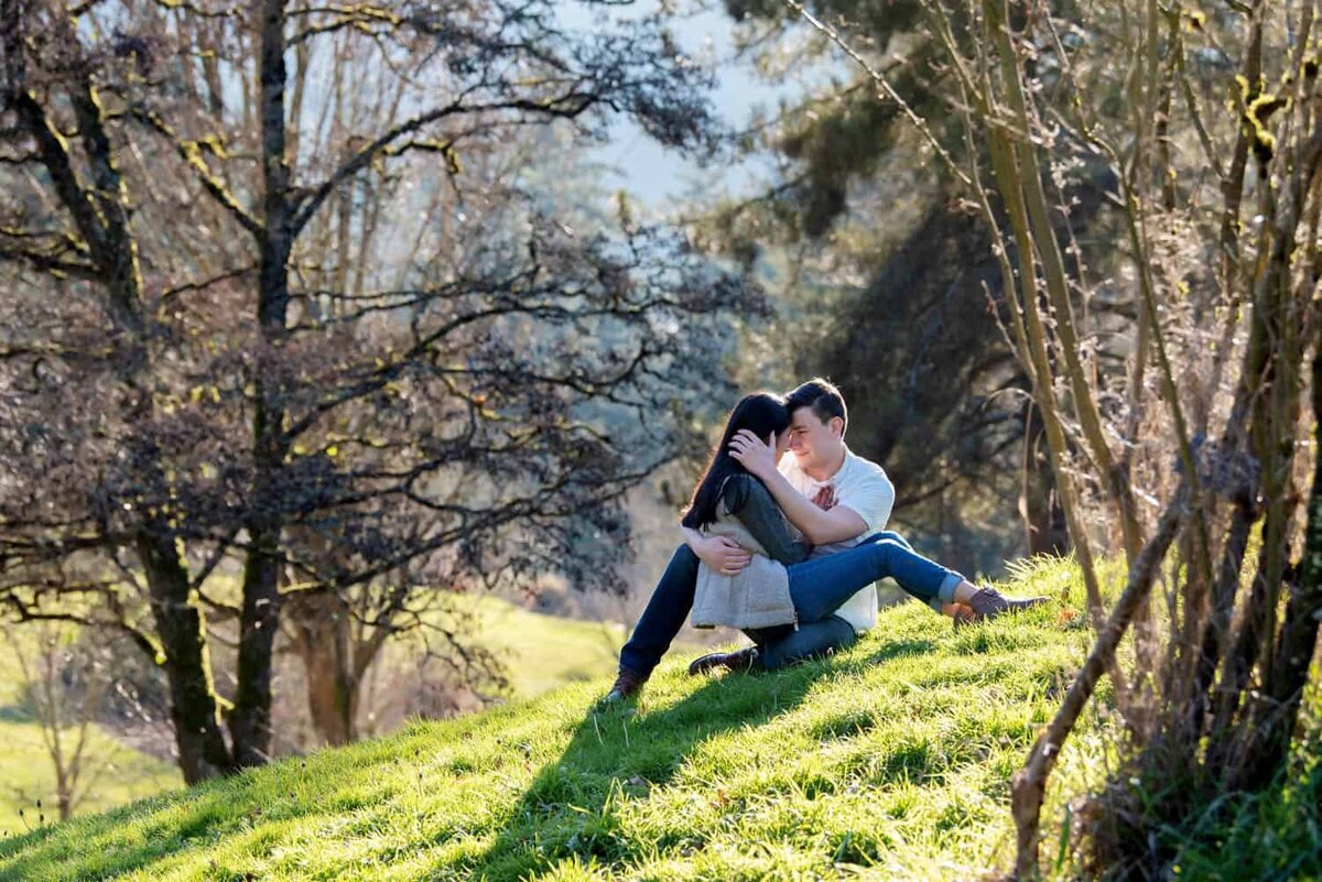 a woman sits on a mans lap in the grass with a beam of sunlight on them