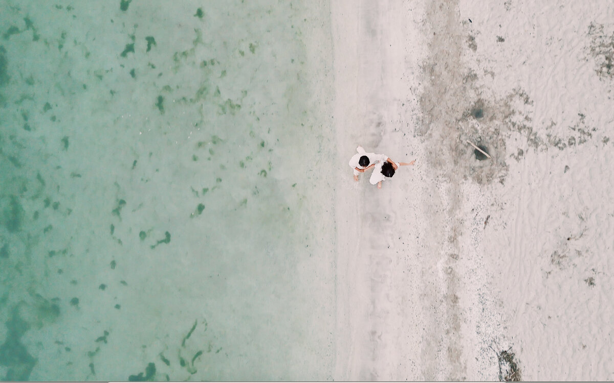 Aerial view of couple walking along a quiet shoreline in soft neutral tones