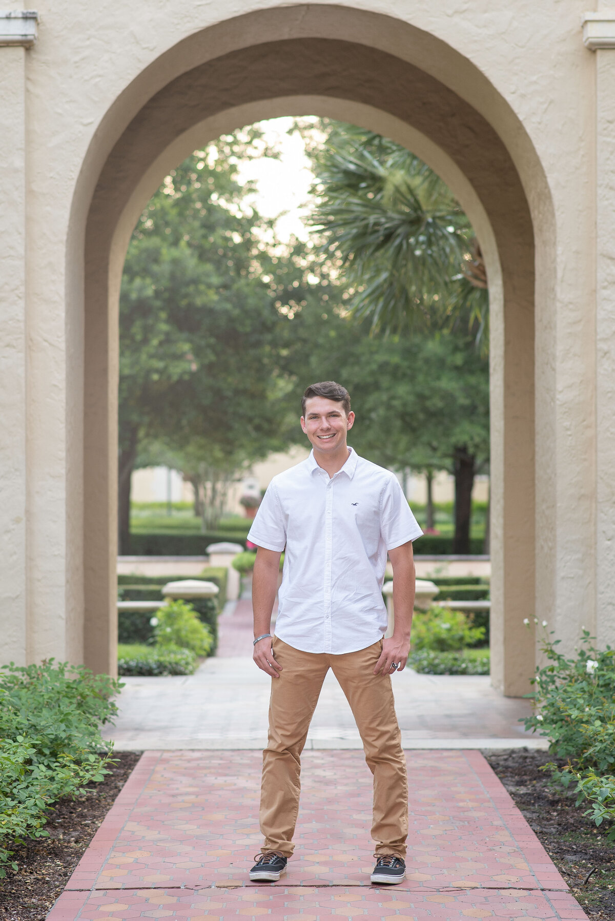 High school senior boy standing on a path in front of a large stone archway smiles at the camera.